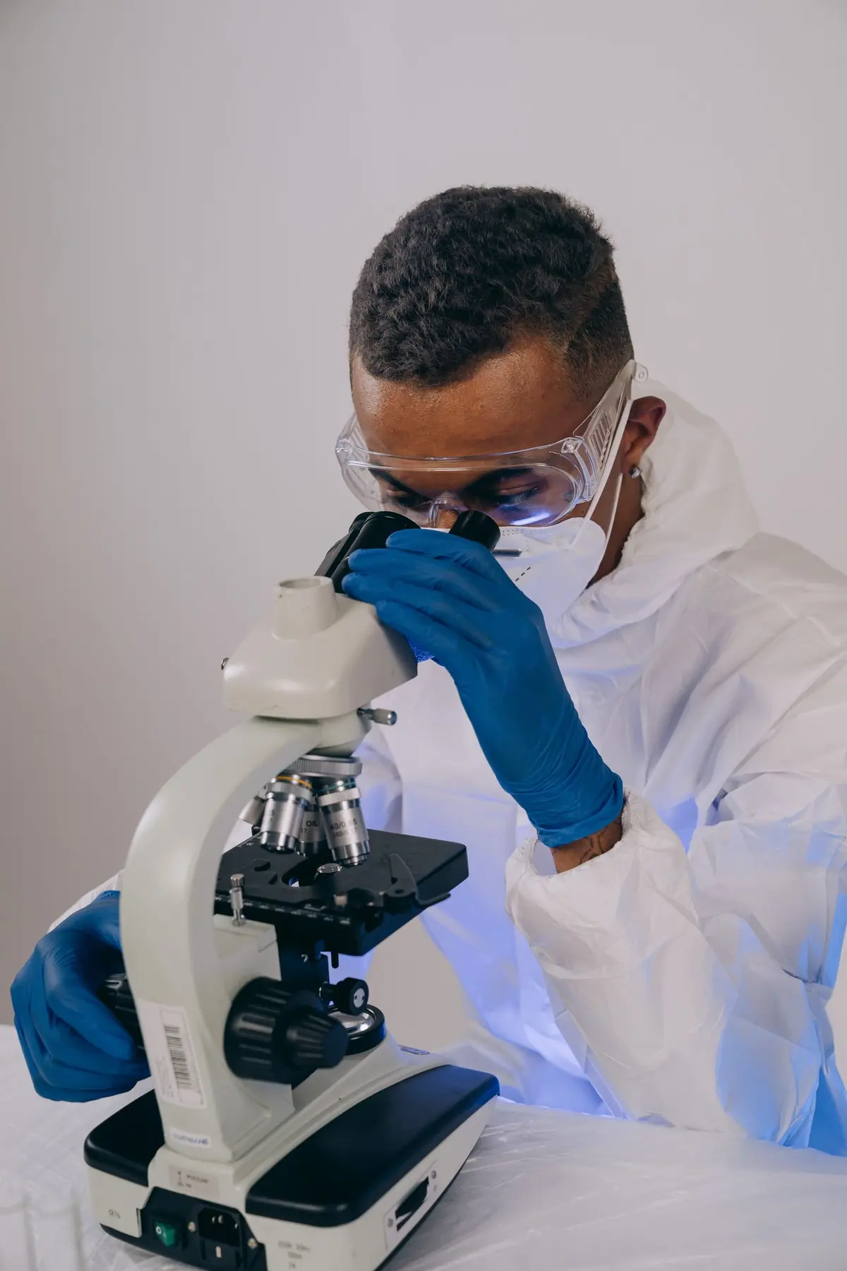 Laboratory technician in protective coveralls, goggles and gloves analysing a sample under a microscope
